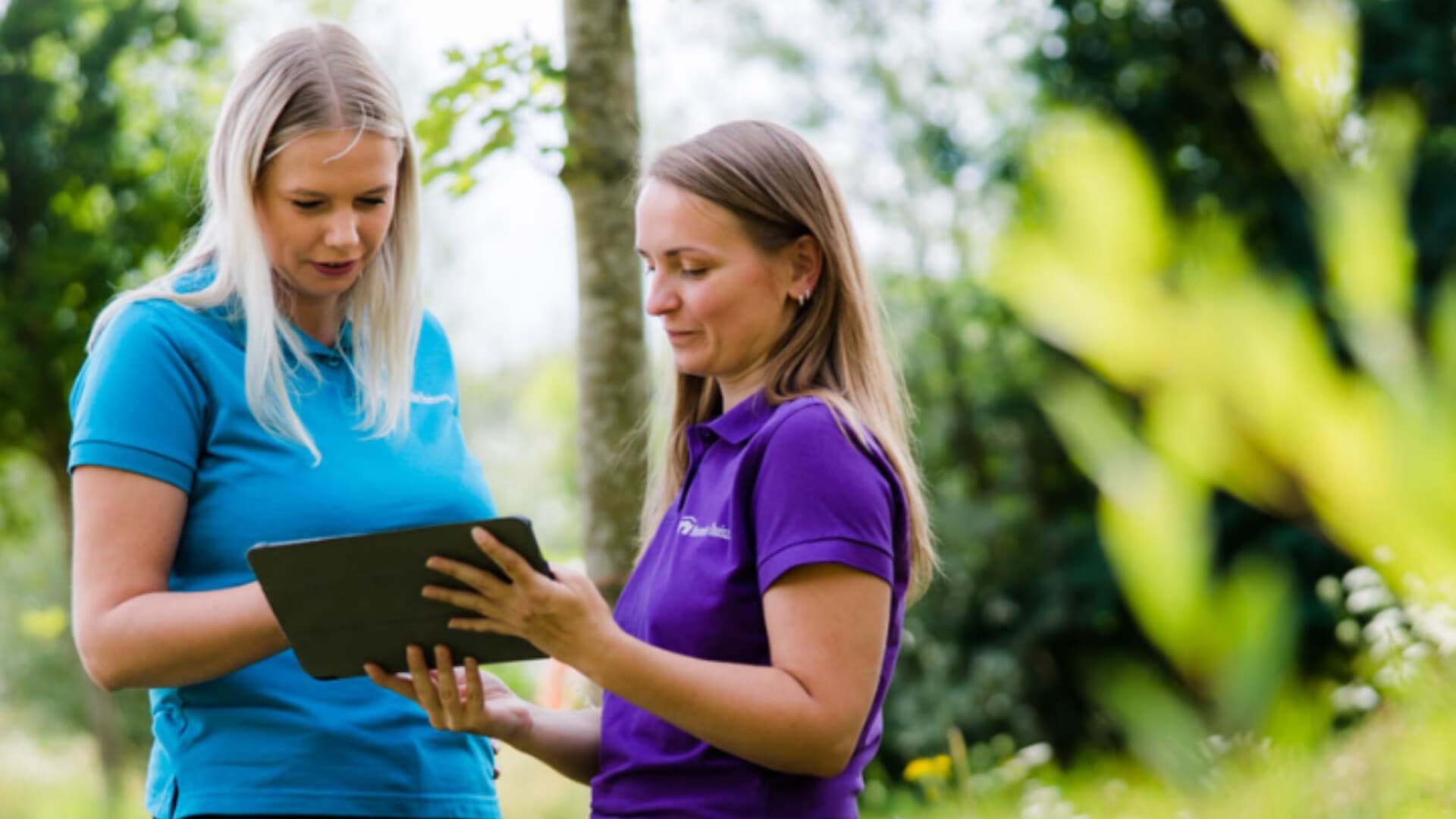 Twee vrouwen kijken naar een Ipad in een groene omgeving. Ze dragen een blauw en paars shirt van MeanderGroep.