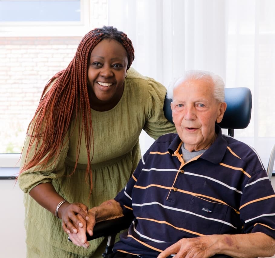 Een glimlachende, jonge vrouw staat achter een oudere man in een rolstoel en houdt zijn hand vast.