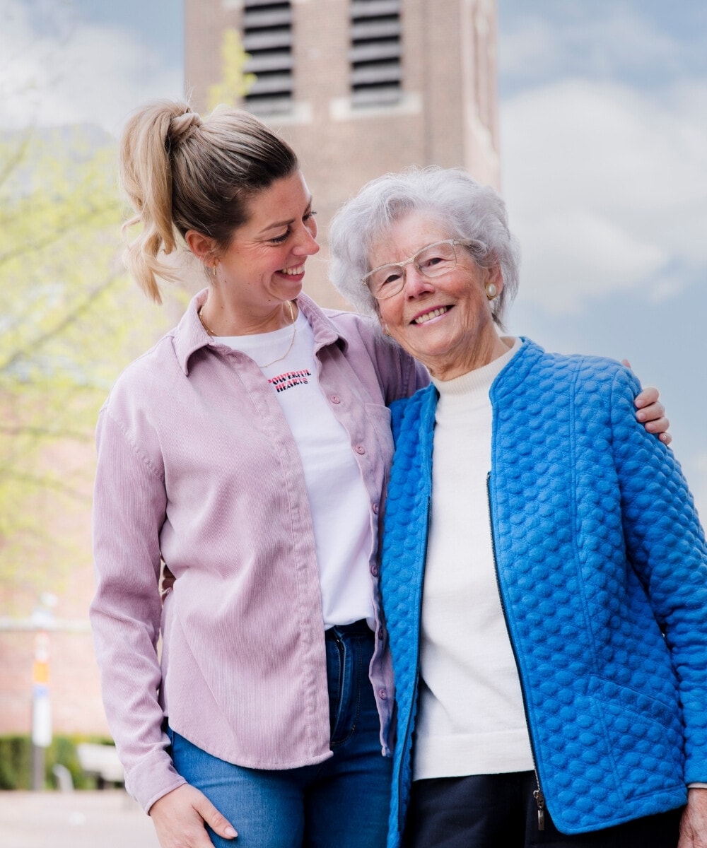 Twee vrouwen, een jongere en een oudere, glimlachen en omarmen elkaar buiten, met een gebouw en bomen op de achtergrond.