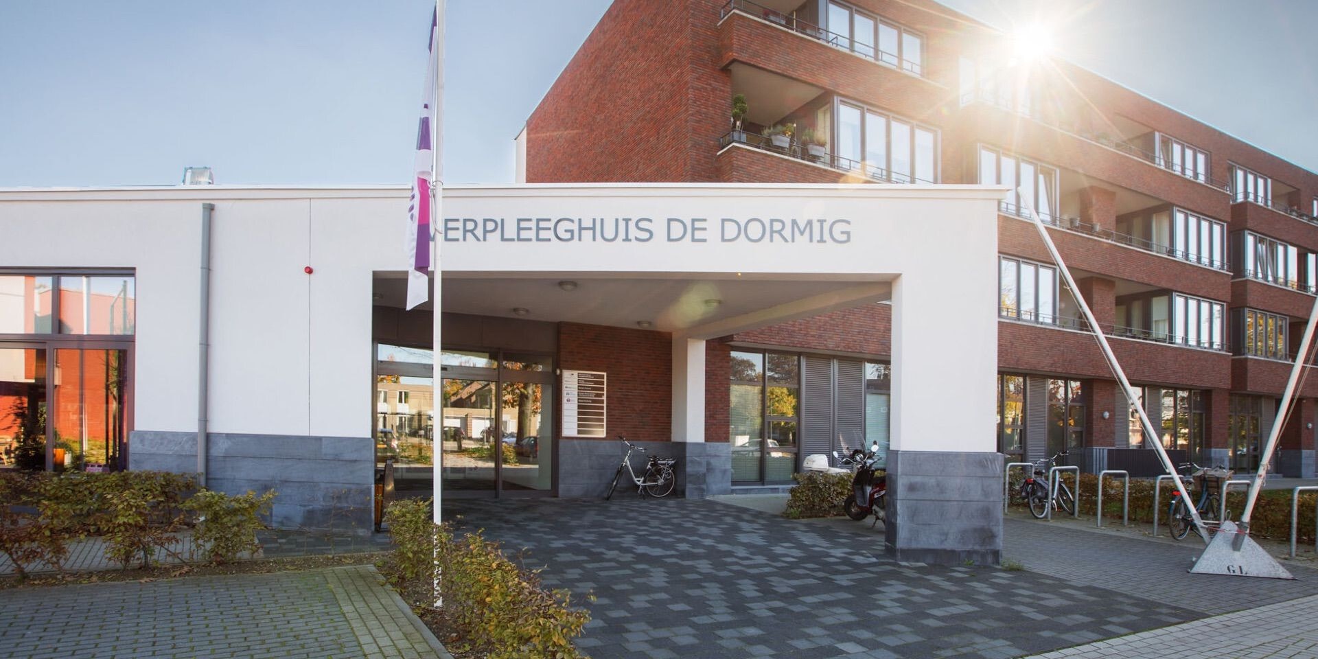 De witte entree van verpleeghuis De Dormig baadt in het zonlicht, met op de achtergrond het moderne bakstenen hoofdgebouw en een parkeerplaats voor fietsen.