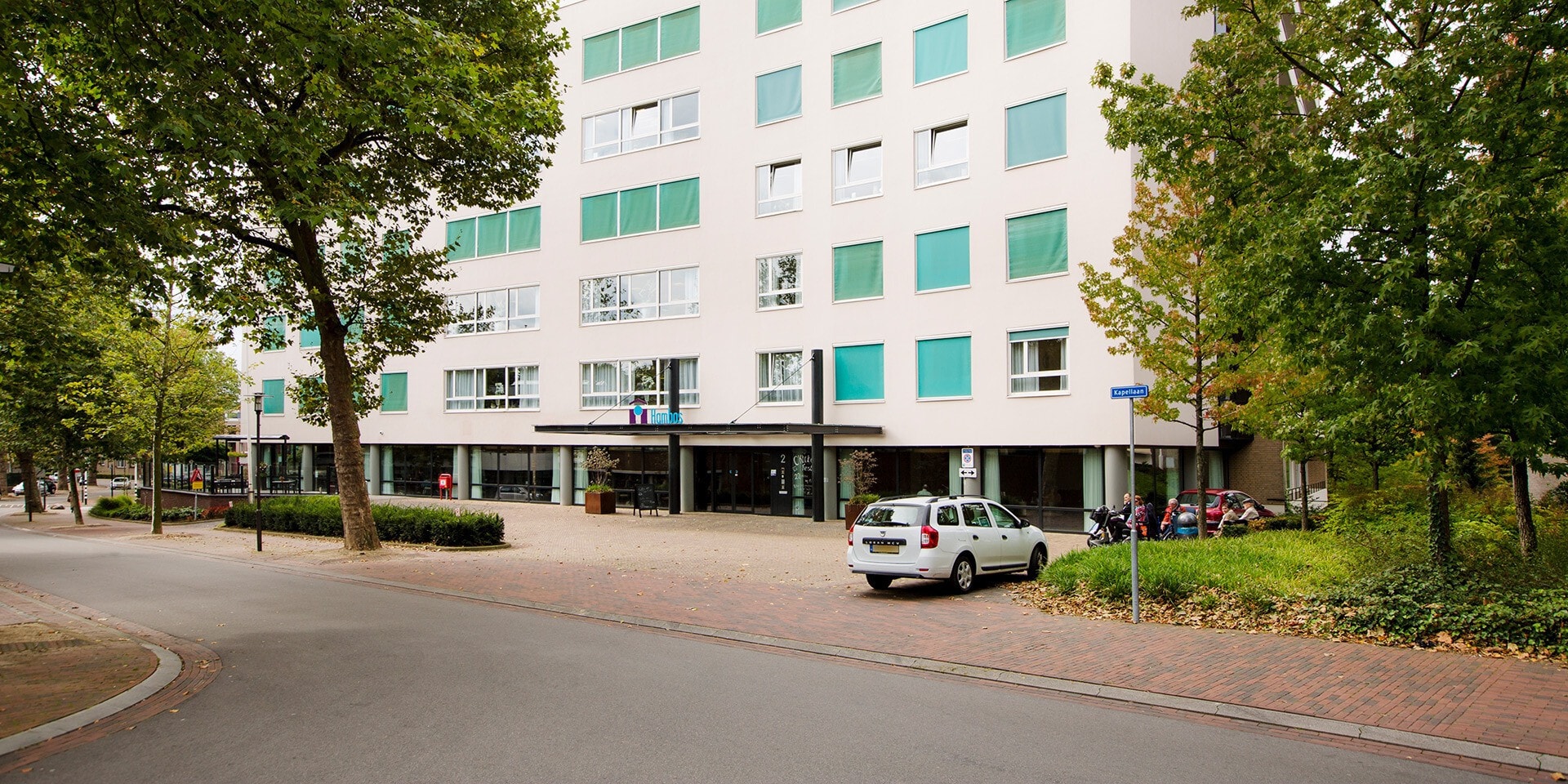 Een modern, wit appartementencomplex met groene zonweringen aan de ramen, gelegen aan een rustige straat met grote bomen en een parkeerplaats voor de deur.