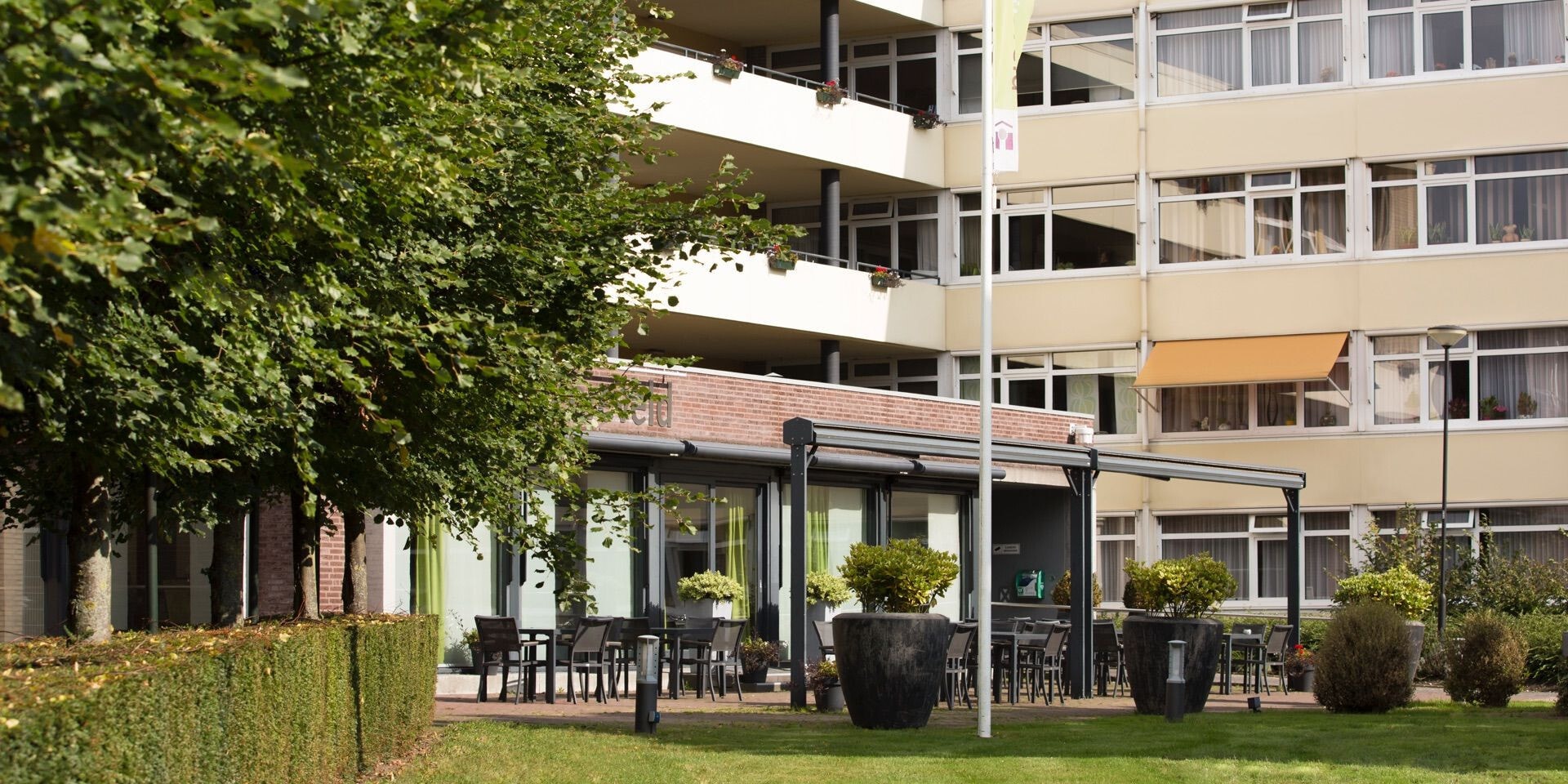 Een overzicht van een verzorgd buitenterras bij een appartementencomplex, voorzien van zwarte terrasmeubels en grote bloempotten, omgeven door groen en bomen.