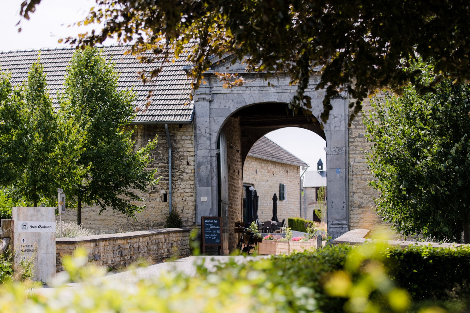 De stenen toegangspoort van de historische Hoeve Overhuizen geeft een doorkijkje naar de binnenplaats met een terras, omgeven door groen en bomen.