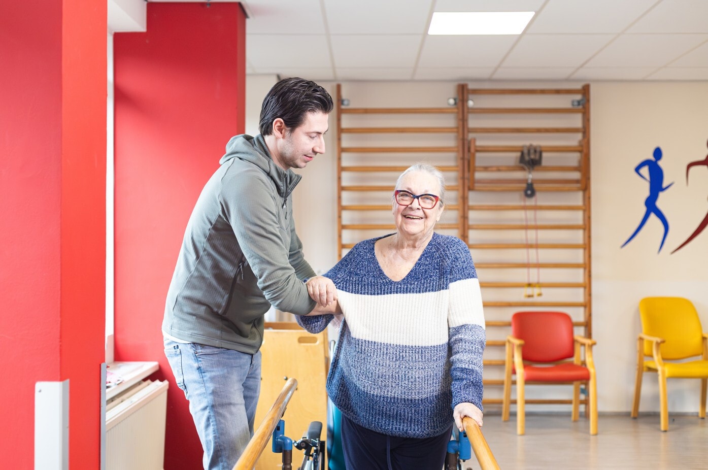 Een lachende oudere vrouw met een bril oefent met lopen tussen twee houten parallelle stangen in een revalidatieruimte. Een jonge man ondersteunt haar door haar arm vast te houden.
