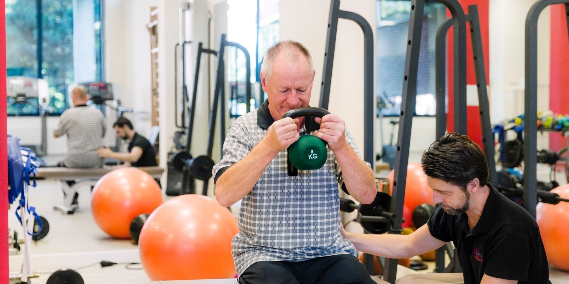 Een oudere man zit op een fitnessbank in een sportschool en houdt een groene kettlebell van 8 kg met beide handen voor zijn borst vast. Een fysiotherapeut met een zwart poloshirt begeleidt hem.