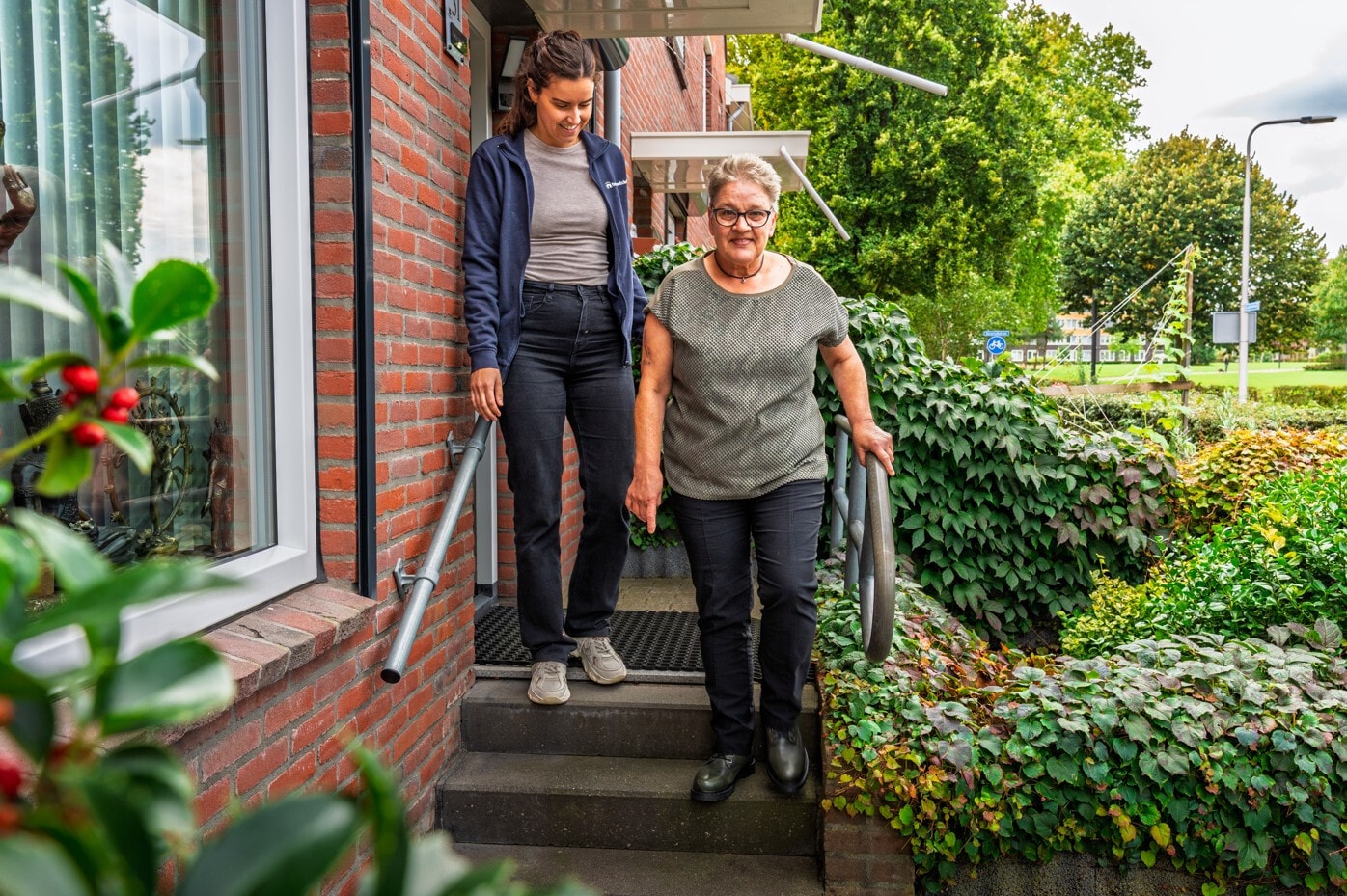 Een jonge vrouw loopt naast een oudere vrouw een stenen trap voor een bakstenen huis af.