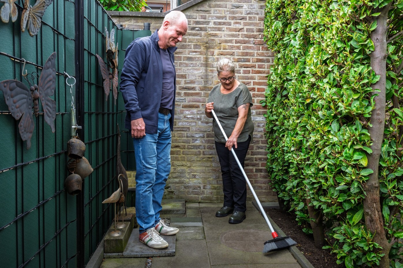 Een vrouw met een bril en een groen shirt veegt met een bezem de tegels van een smal pad in een tuin. Naast haar staat een man in een blauw vest en spijkerbroek die glimlachend toekijkt.