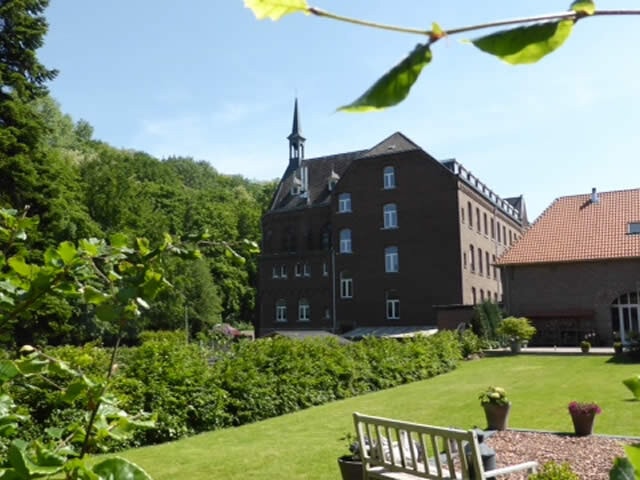 Een historisch bakstenen gebouw met een karakteristiek torentje, omgeven door een uitgestrekte groene tuin met een gazon en dichte bebossing op de achtergrond.