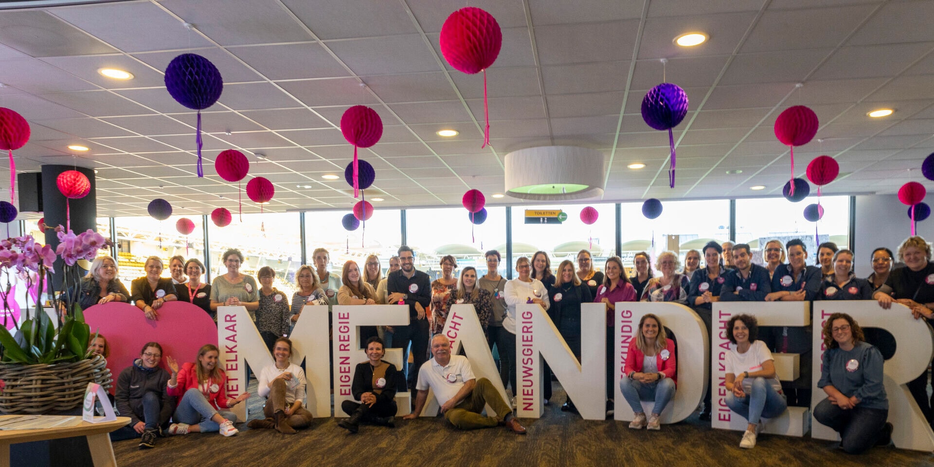 Een grote groep mensen, voornamelijk vrouwen, poseert lachend achter en rondom grote witte letters die het woord 'MEANDER' spellen. De ruimte is versierd met roze en paarse lampionnen.