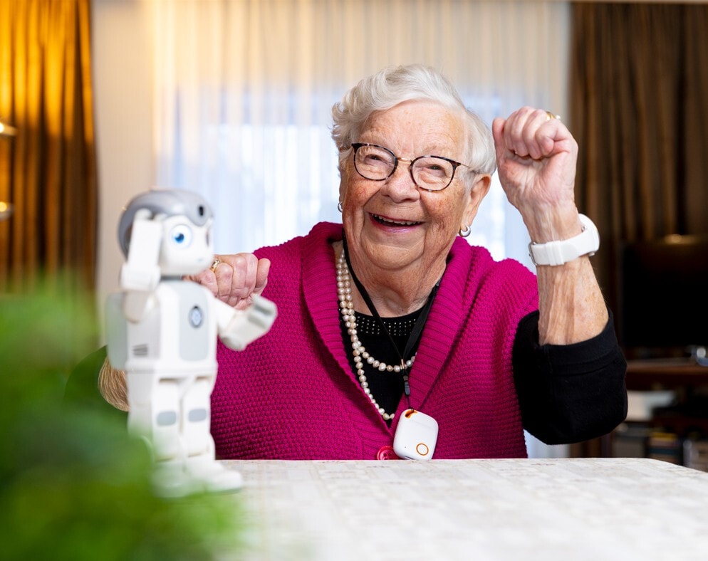Een lachende oudere vrouw met een bril en een felroze vest imiteert de bewegingen van een kleine, witte robot die voor haar op tafel staat.