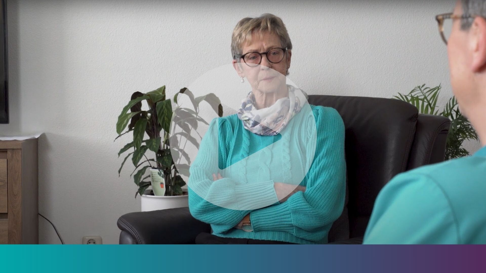 Een oudere vrouw met kort blond haar en een bril zit met haar armen over elkaar in een zwarte lederen fauteuil. Over de afbeelding heen staat een grote afspeelknop.