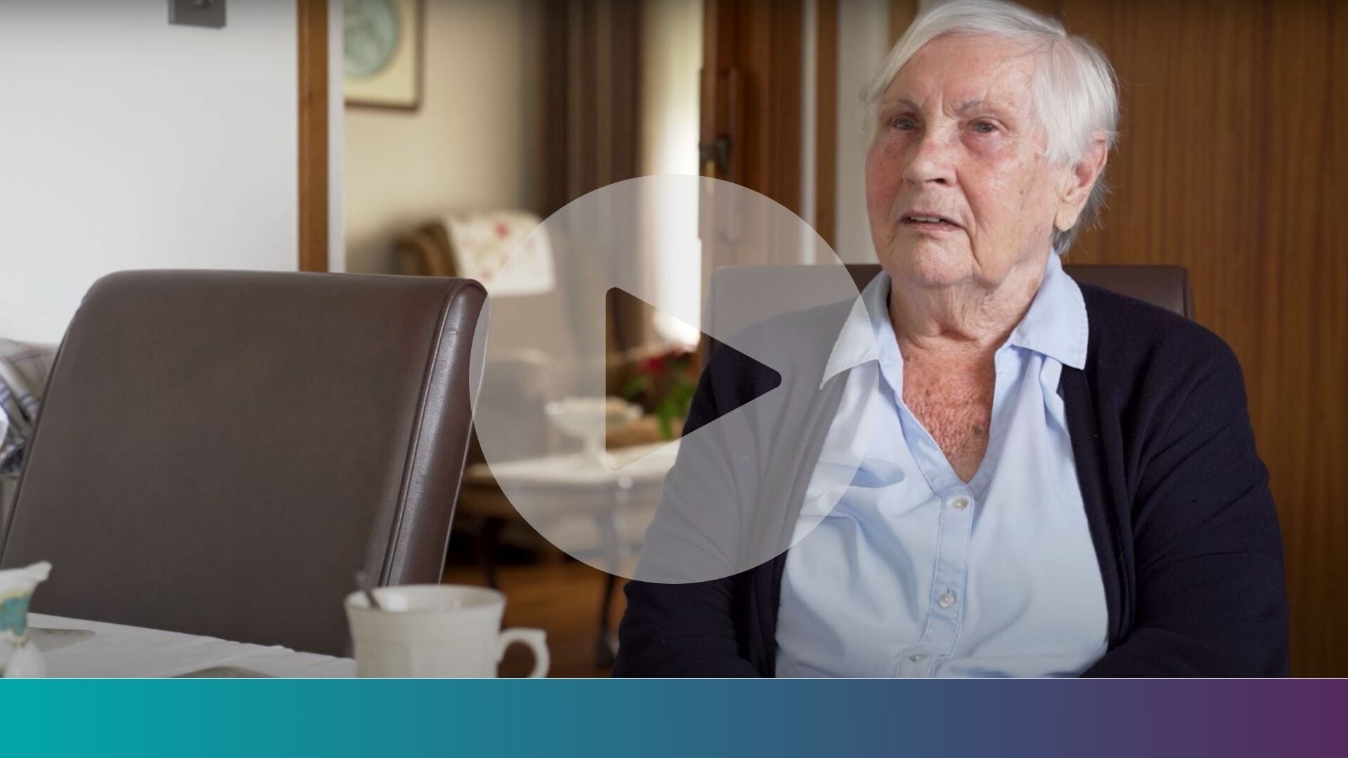 Een oudere vrouw zit aan een eettafel in een huiselijke omgeving. Over de afbeelding heen staat een grote afspeelknop.