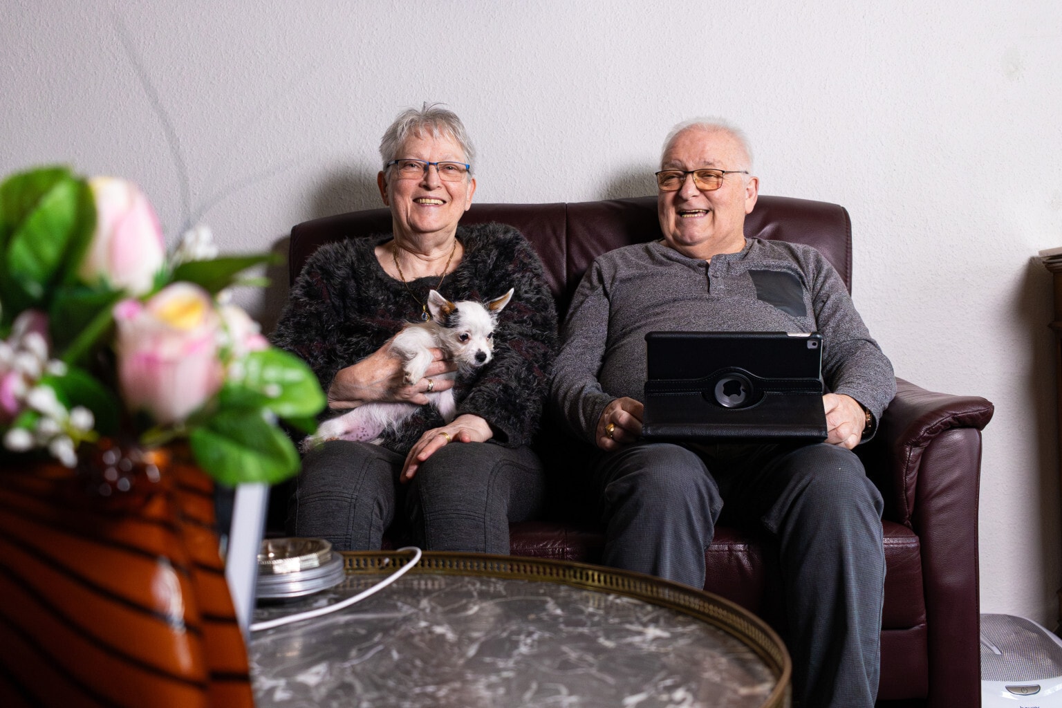 Lachende oudere man en vrouw zitten op een bruine bank. De vrouw heeft een klein hondje op haar schoot.