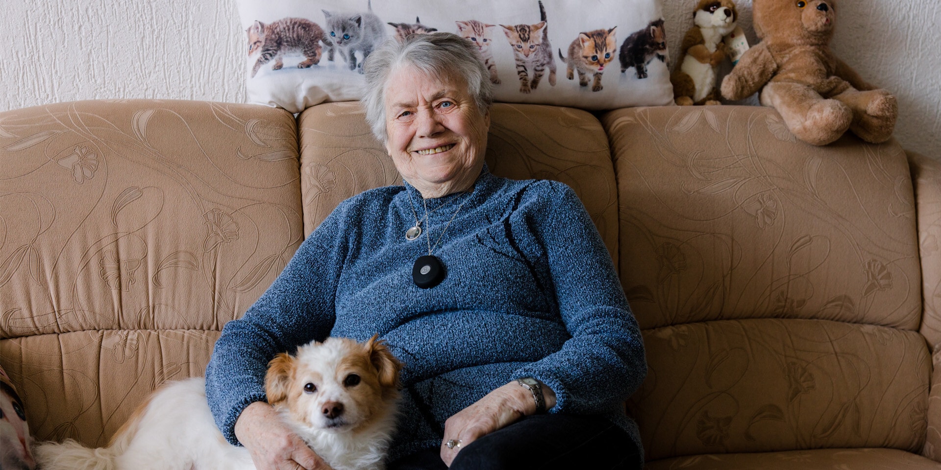Een lachende oudere vrouw zit op een bruine bank, op haar schoot ligt een hondje.