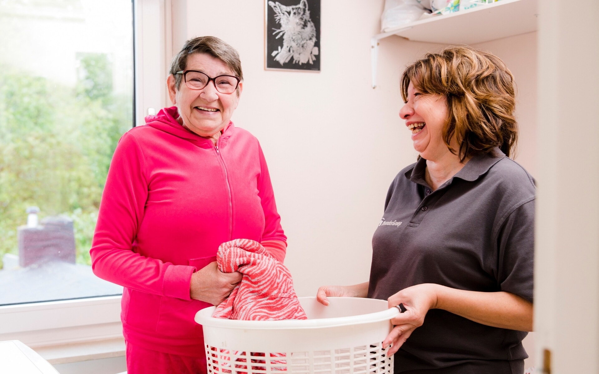 Een blije oudere cliënt en een zorgverlener lachen samen terwijl ze de was opvouwen. De oudere vrouw draagt een roze trainingspak. De zorgverlener houdt een witte wasmand vast en draagt een zwarte polo van MeanderGroep.