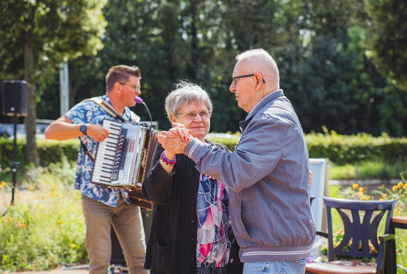 Oudere vrouw en man dansen buiten met elkaar. Achter hen staat een man voor een microfoon met een accordeon in zijn handen.