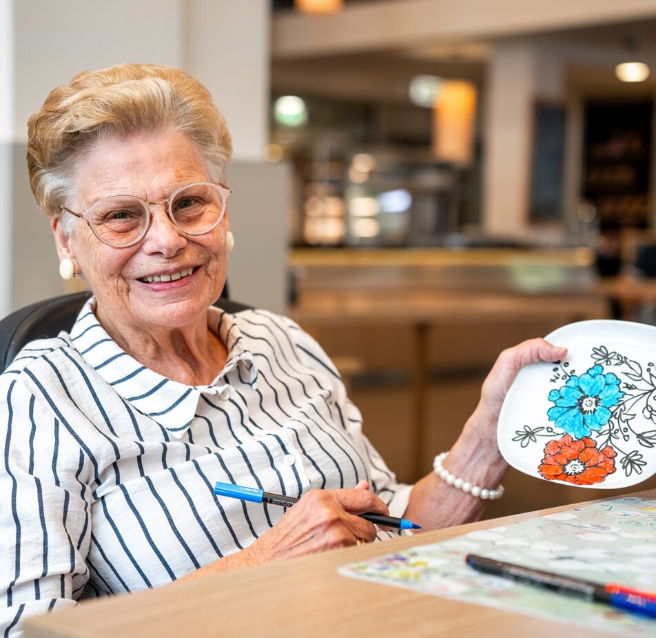Een glimlachende oudere vrouw met een bril toont trots een wit bordje dat ze heeft versierd met blauwe en oranje bloemen. Ze zit aan een tafel met een blauwe viltstift in haar hand.