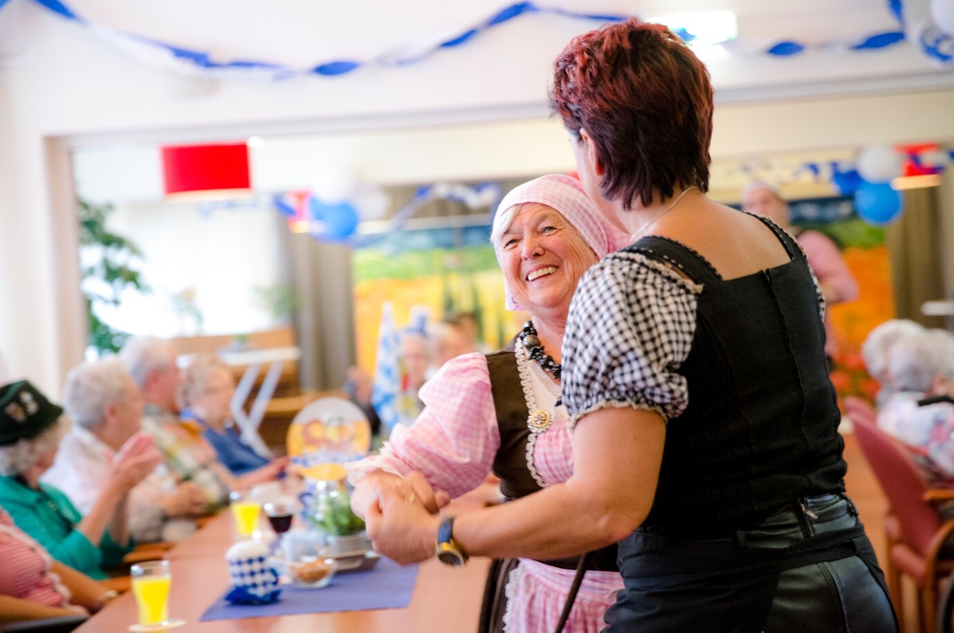 Een lachende vrouw in tiroler klederdracht danst tijdens een feestelijke bijeenkomst, terwijl andere gasten aan lange tafels toekijken en meeklappen.