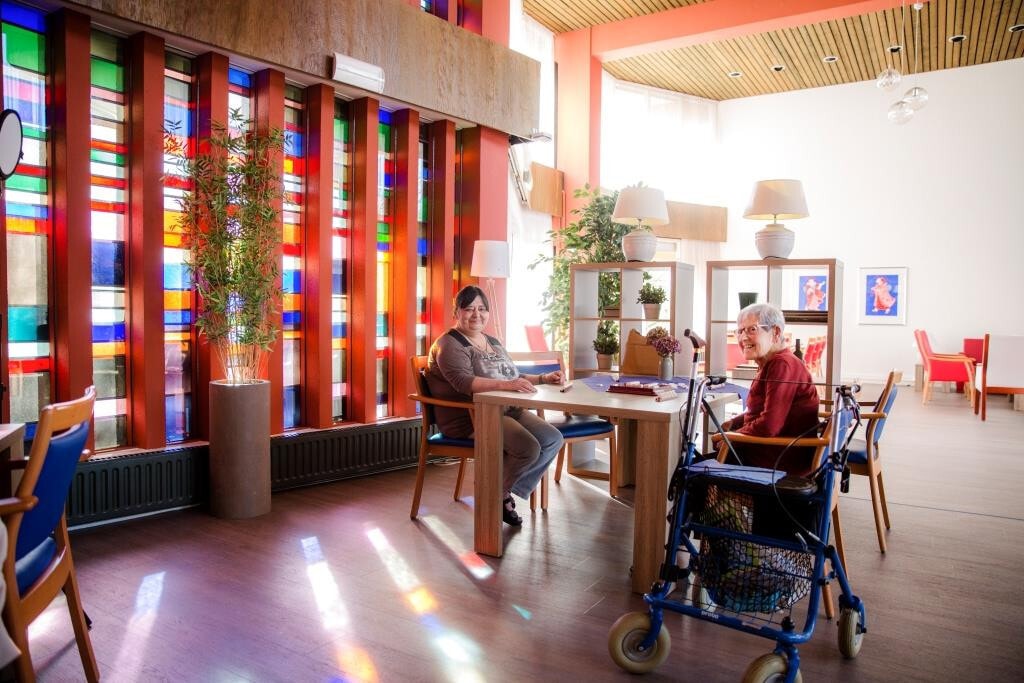 Twee vrouwen zitten gezellig aan een tafel in een lichte, kleurrijke ruimte met glas-in-loodramen die voor een sfeervolle lichtinval op de vloer zorgen.