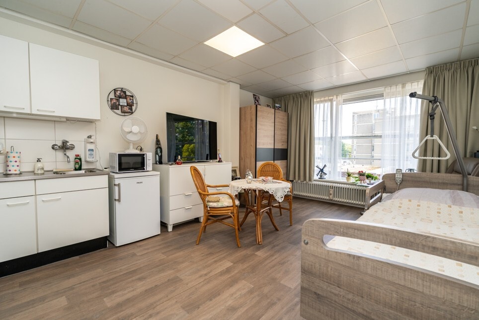 Een overzicht van een gecombineerde woon- en slaapkamer in een zorginstelling, compleet met een bed, een klein keukenblok en een zithoekje bij het raam.