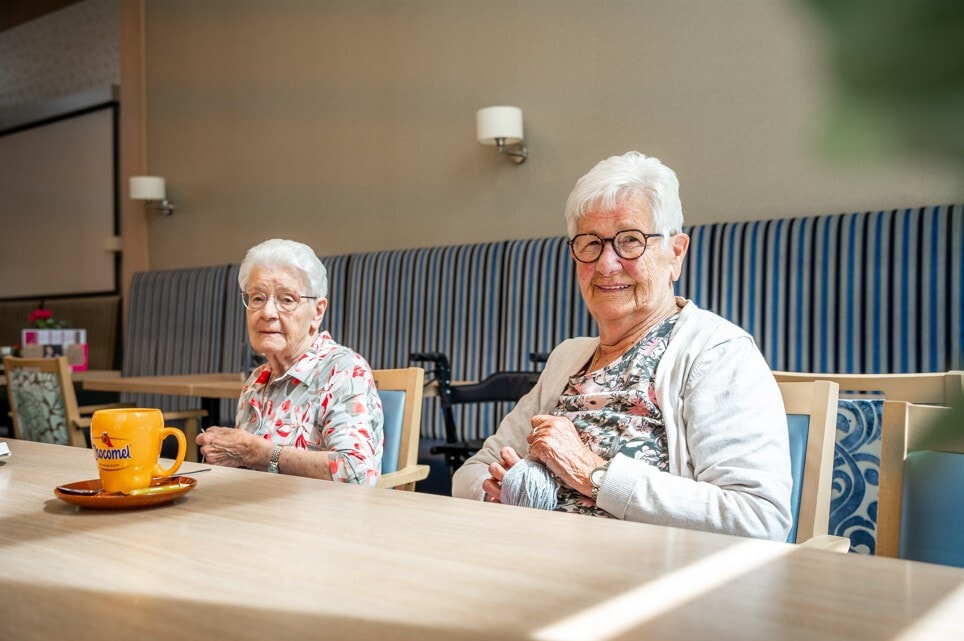 Twee oudere dames zitten ontspannen aan een tafel in een gemeenschappelijke ruimte; de een is bezig met handwerken terwijl er een kopje Chocomel op tafel staat.