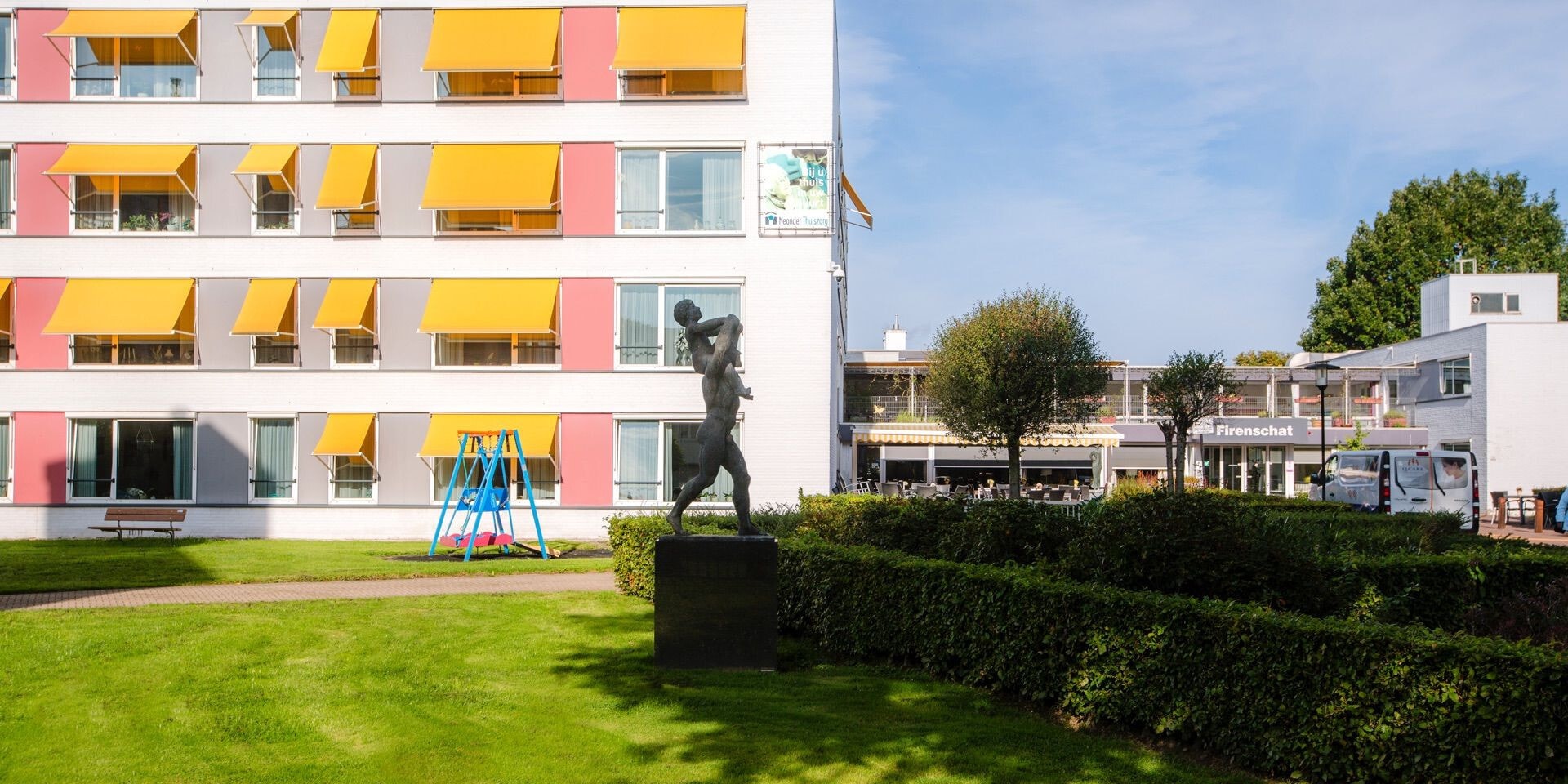 Een modern woon- en zorgcomplex met opvallende gele zonneschermen aan de gevel, gelegen aan een groen gazon met een standbeeld en een verzorgde tuin.
