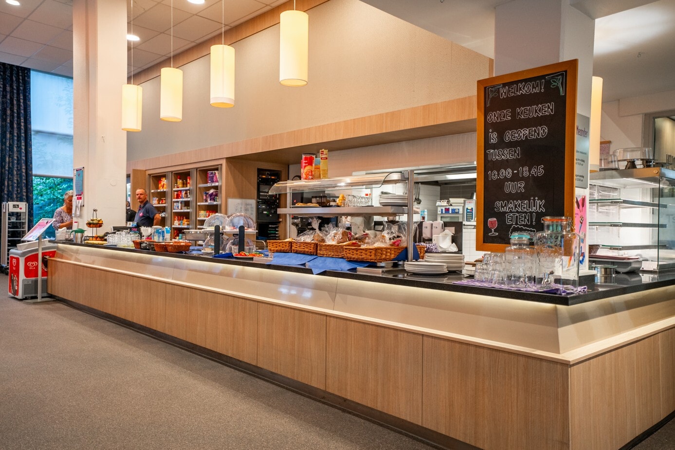 Een modern buffet- en keukengedeelte in een zorginstelling met een lange houten balie, waarop diverse broodjes en drankjes gepresenteerd worden onder sfeervolle hanglampen.