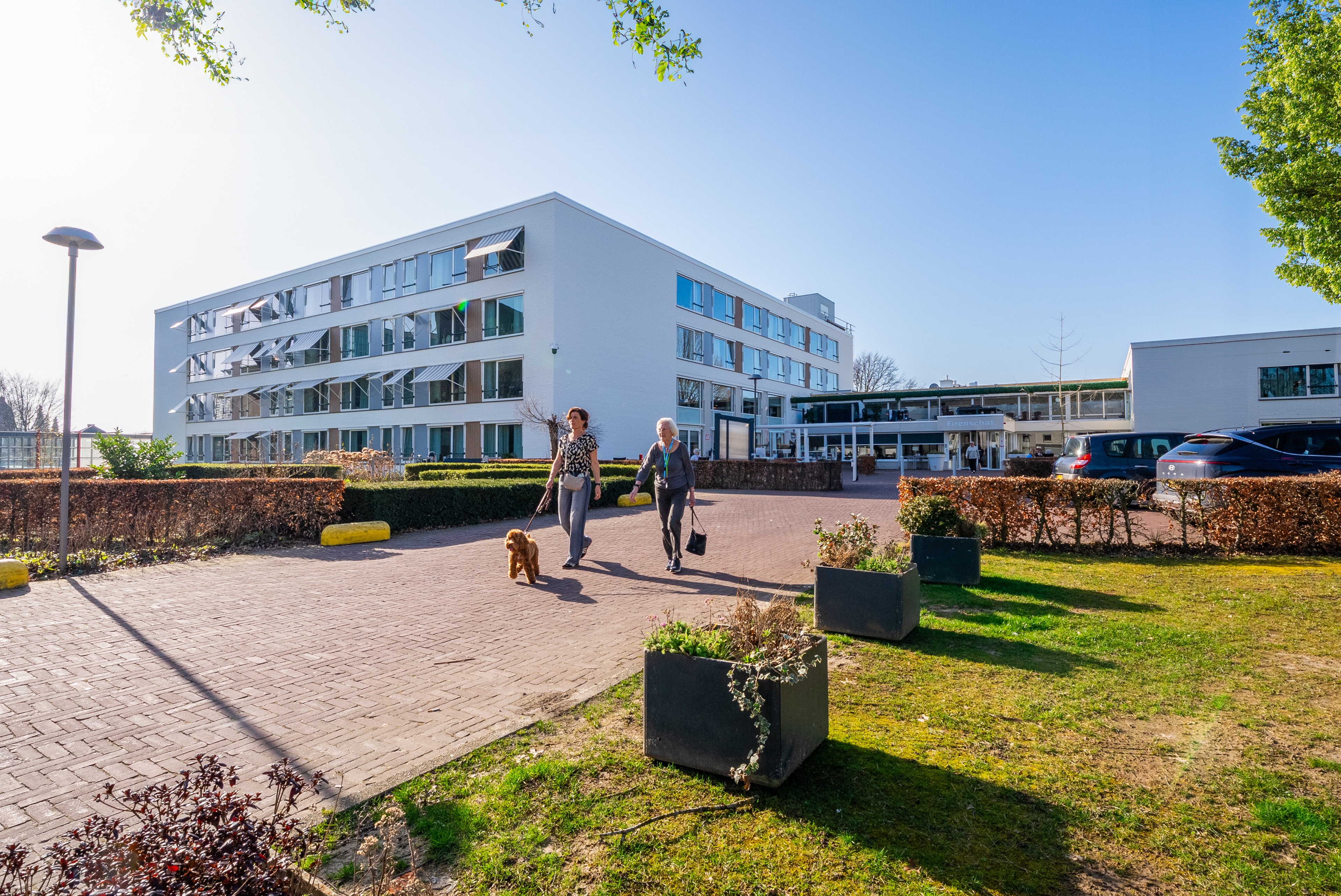 Twee vrouwen wandelen met een hond over een geplaveid pad bij een modern, wit zorgcomplex op een zonnige dag met een strakblauwe lucht.