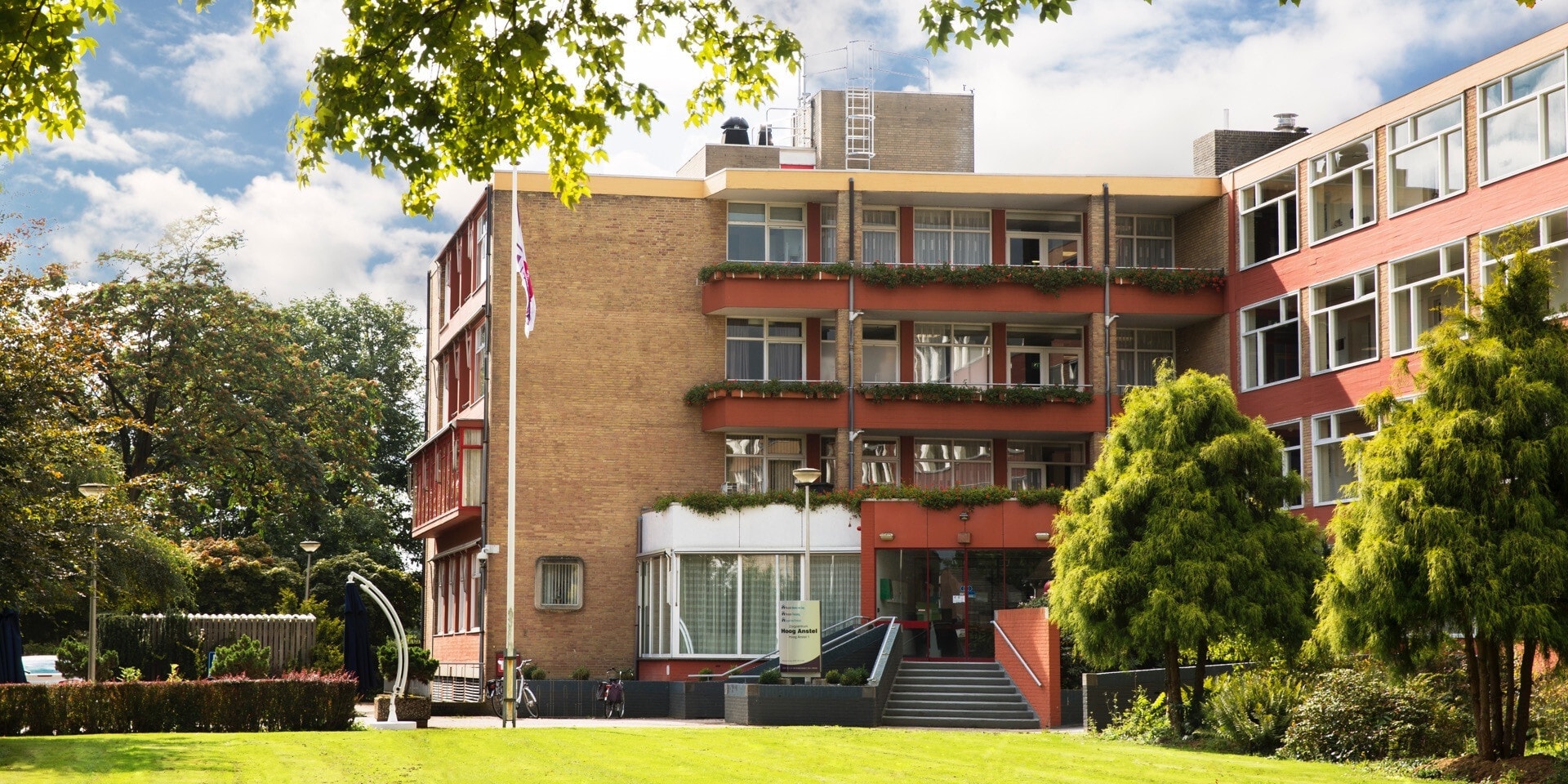 Een zonnig overzicht van een bakstenen zorgcomplex met rode accenten en balkons vol bloembakken, omgeven door een uitgestrekt gazon en volwassen bomen.