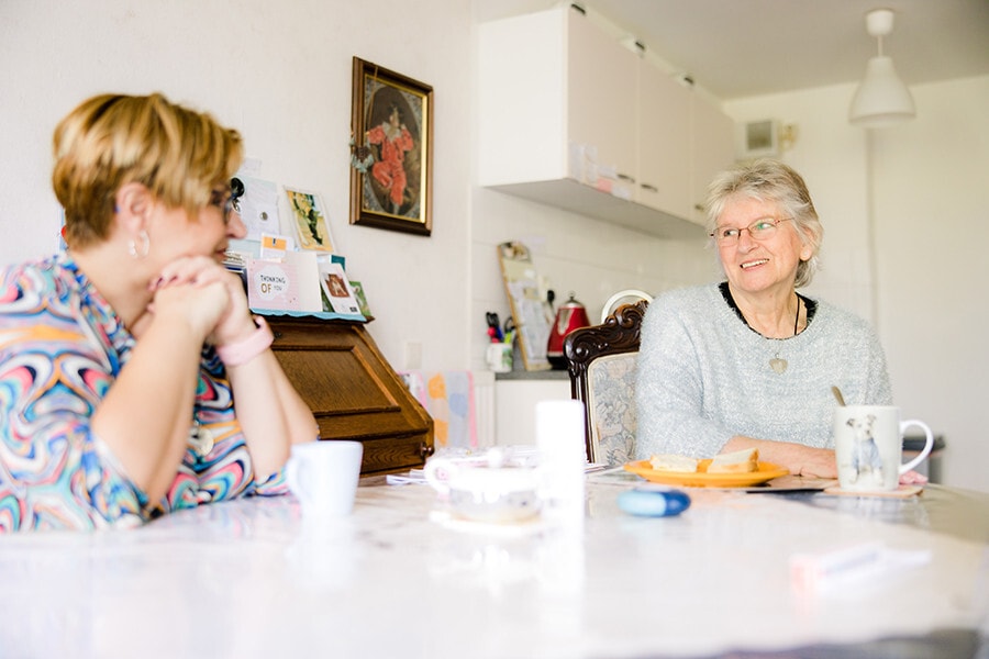 Een oudere vrouw met grijs haar en een bril zit glimlachend aan een witte eettafel in een lichte keuken. Tegenover haar zit een vrouw die aandachtig naar haar luistert.