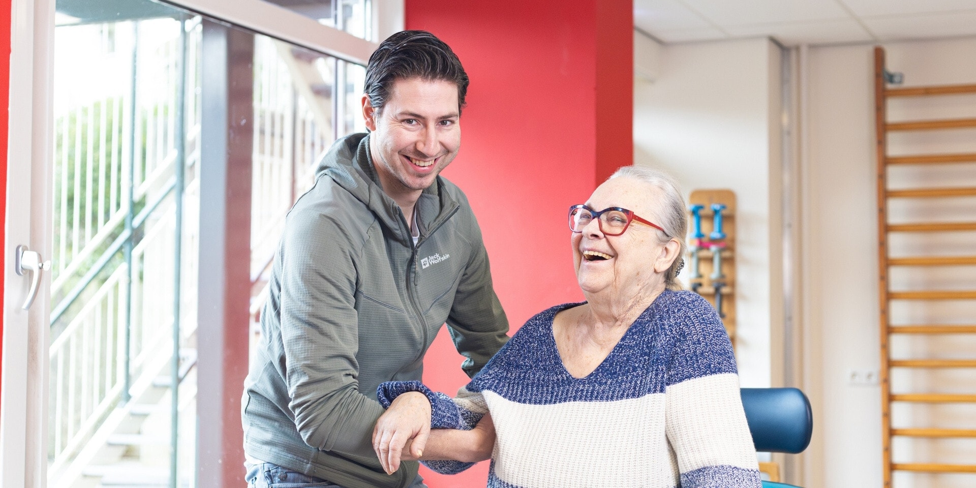 Een jonge mannelijke begeleider ondersteunt een lachende oudere vrouw tijdens een revalidatiesessie.