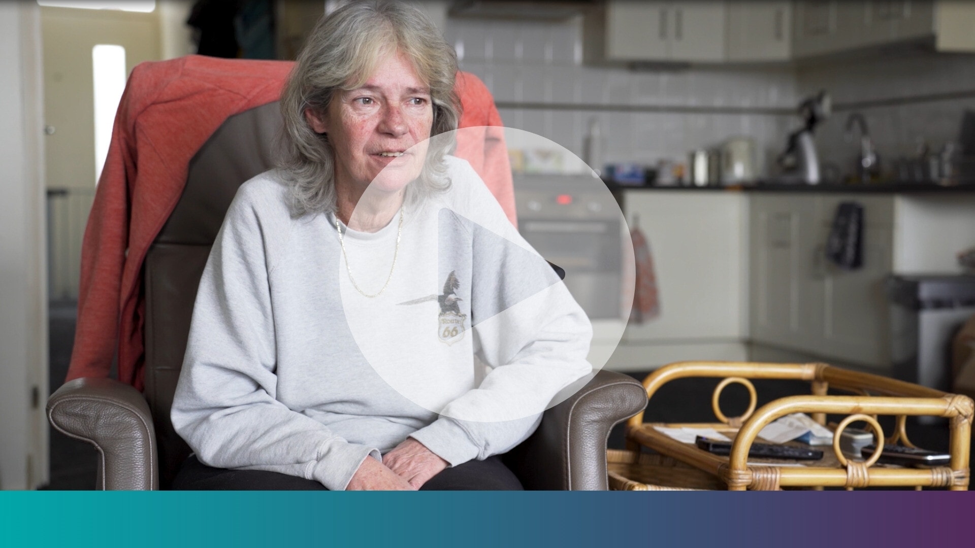 Een vrouw zit in een stoel, met een keuken op de achtergrond en een afspeelknop in het midden van het beeld.
