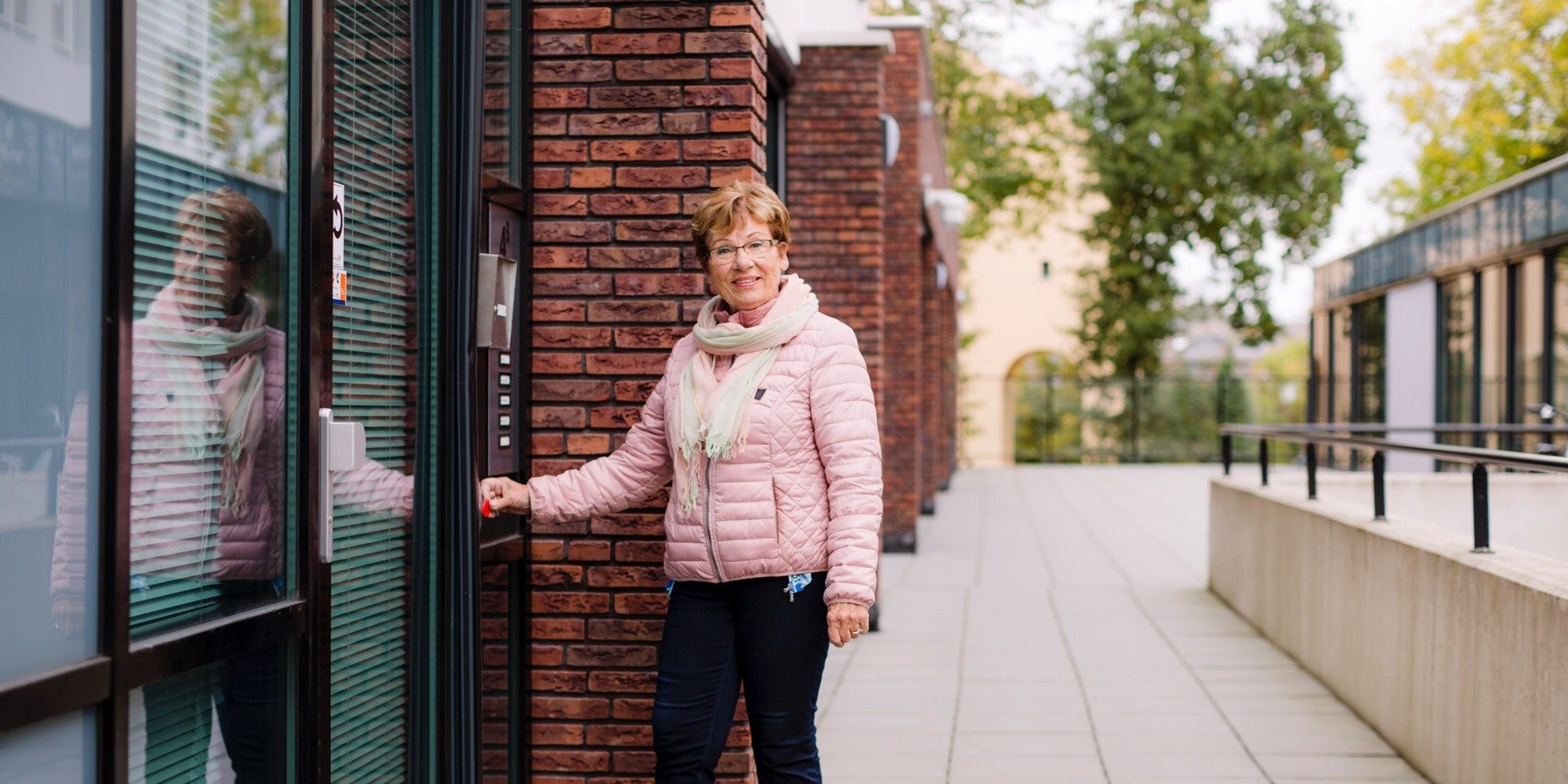Een lachende vrouw met een roze jas staat buiten voor een toegangsdeur van een modern bakstenen gebouw.