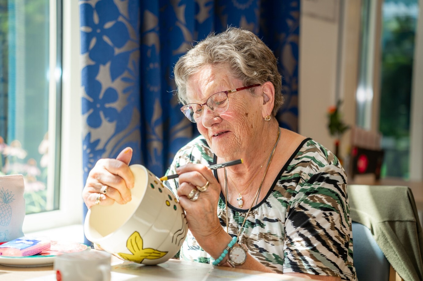 Een oudere vrouw met een bril is geconcentreerd bezig met het versieren van een witte keramieken schaal met een viltstift.