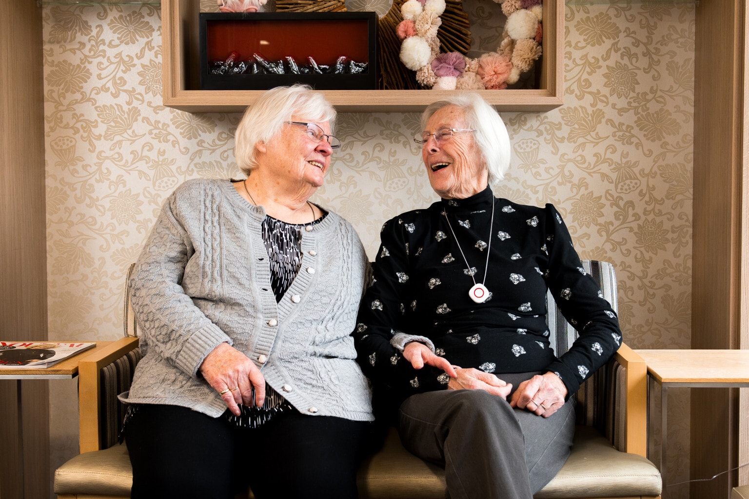 Twee oudere dames zitten naast elkaar op een bankje en hebben een vrolijk gesprek. Ze lachen naar elkaar in een huiselijke omgeving.