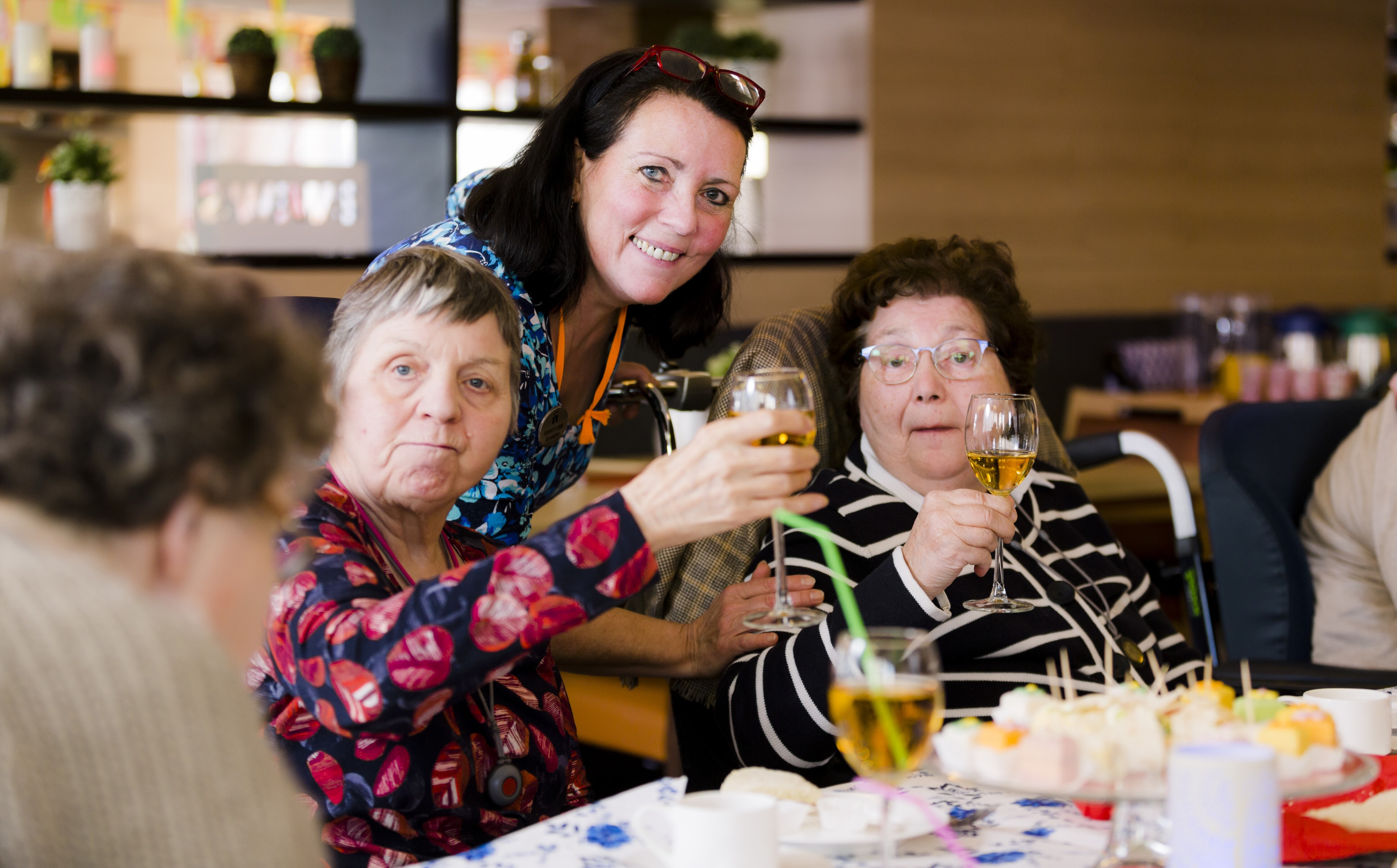 Een groep oudere bewoners, vergezeld door een medewerkster, heffen hun glas voor een proost aan een mooi gedekte tafel.