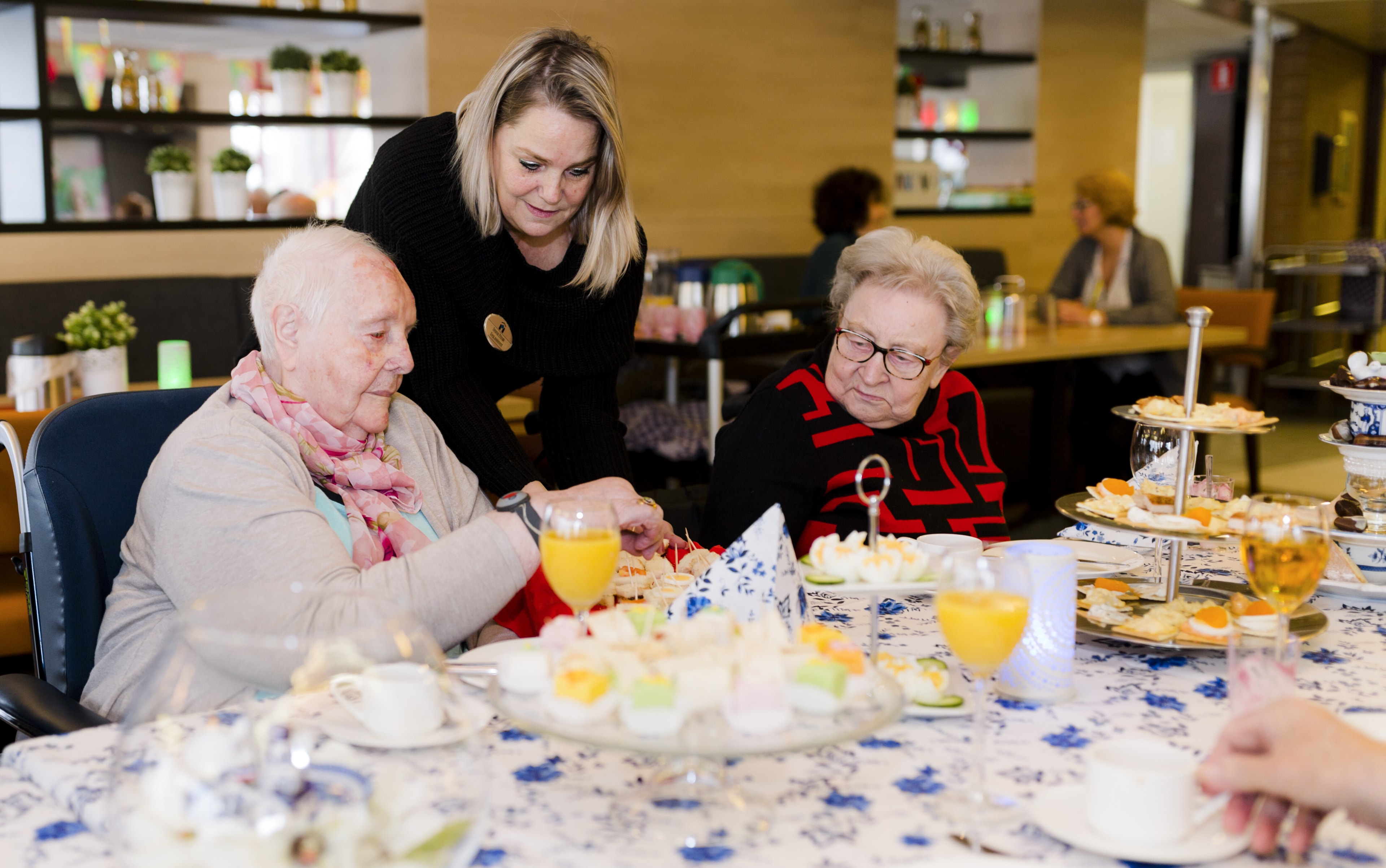 Een medewerkster helpt twee oudere dames aan een feestelijk gedekte tafel met hapjes en drankjes in een gezamenlijke ruimte.