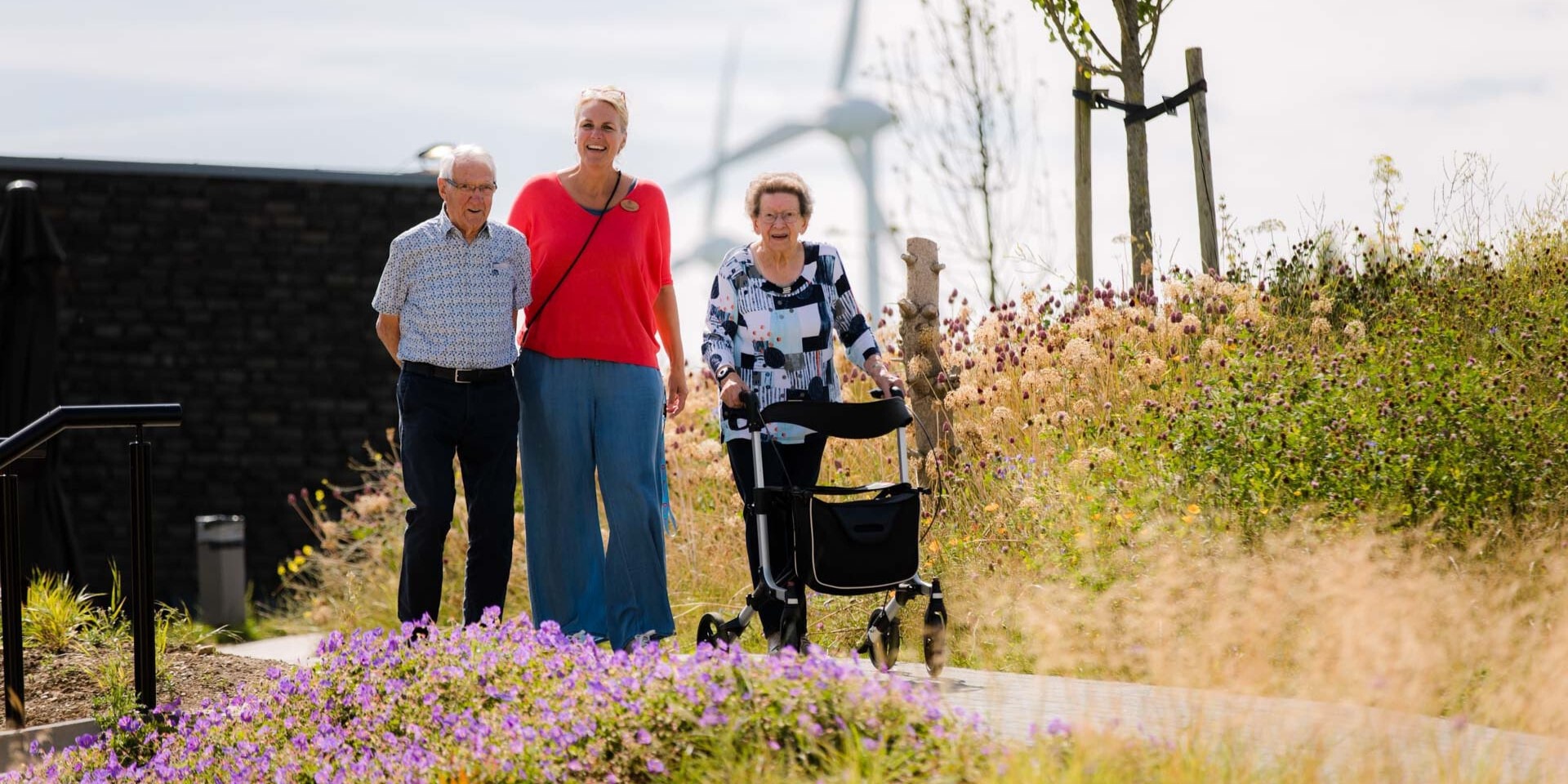 Een jongere vrouw loopt met een oudere man en een oudere vrouw met een rollator door een bloemrijke tuin.
