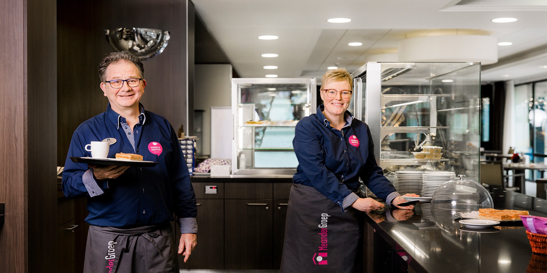 Twee medewerkers van de MeanderGroep, een man en een vrouw, staan glimlachend in een horecagelegenheid.