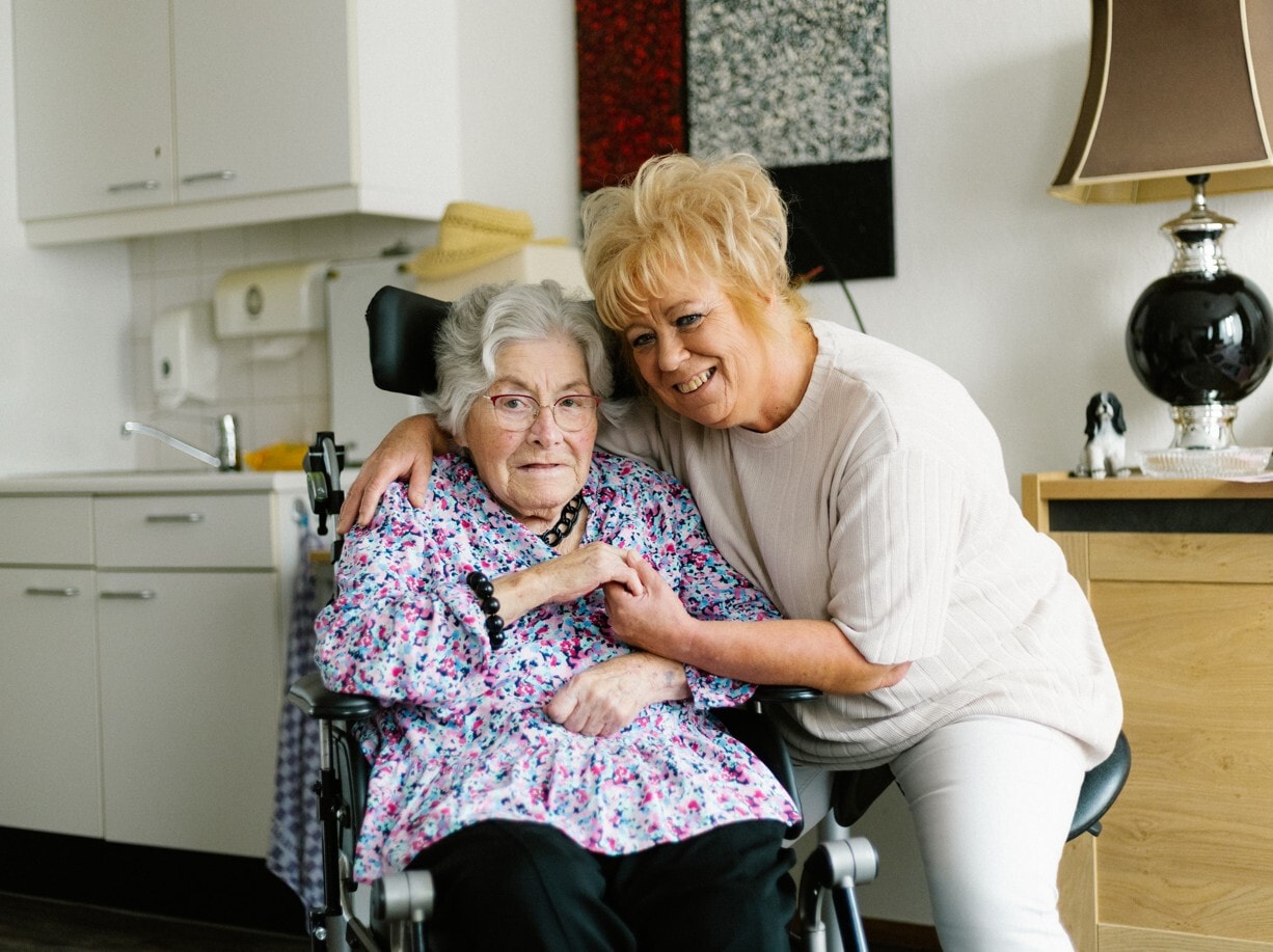 Glimlachende vrouw met blond haar omhelst een oudere vrouw in een rolstoel.