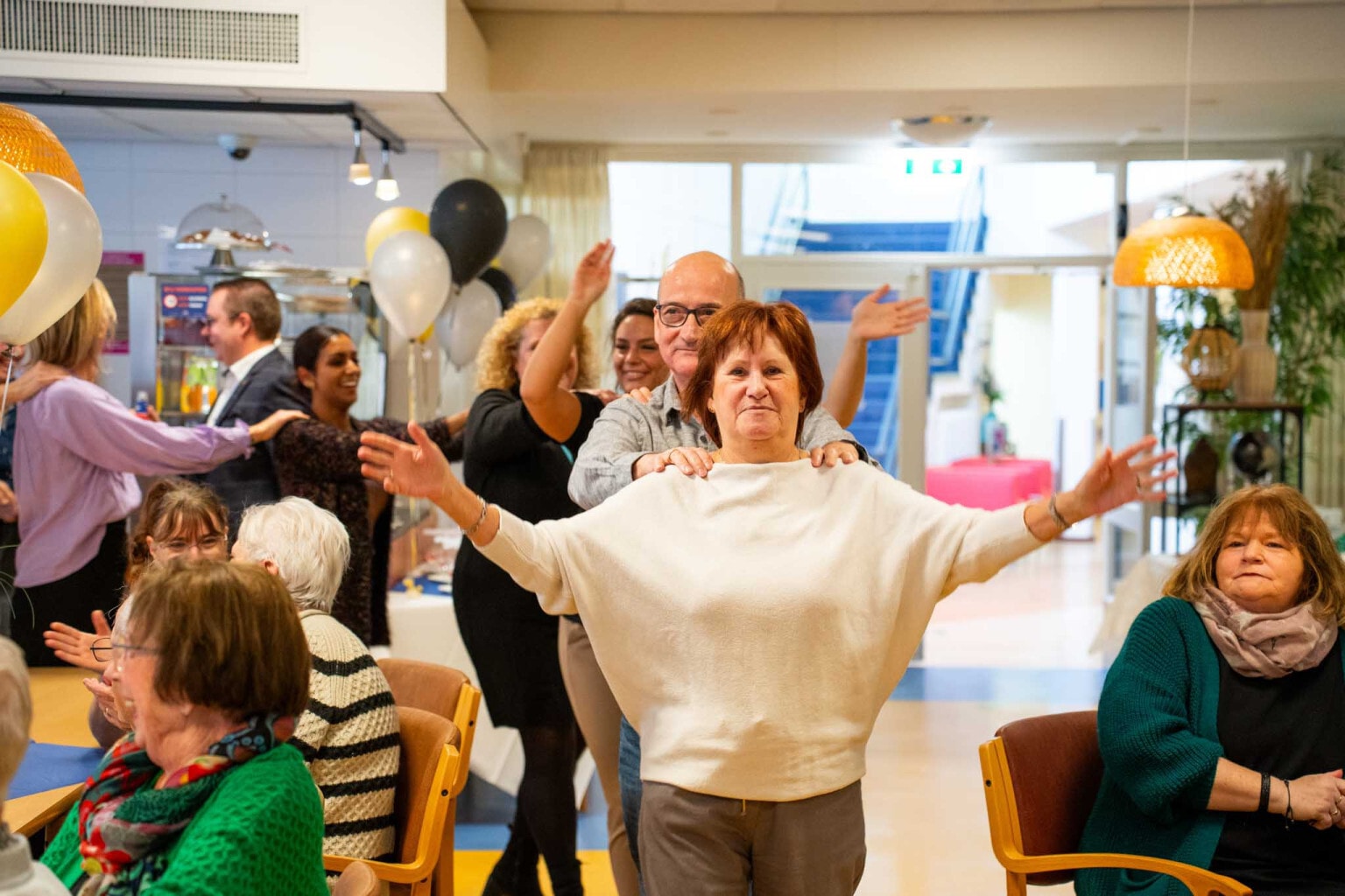 Een feestelijke bijeenkomst in een binnenruimte waarbij een groep mensen een polonaise vormt. Op de stoelen zitten meerdere mensen die lachen en klappen.