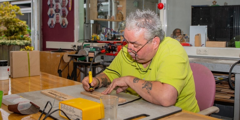 Een man zit geconcentreerd aan een tafel in een werkplaats. Met een brandpen schrijft hij op een houten plankje.