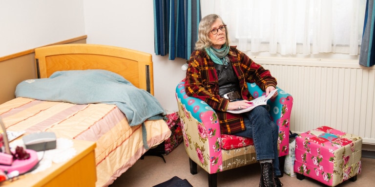 Een vrouw met een bril zit in een kleurrijke, gebloemde stoel naast een bed in een slaapkamer.