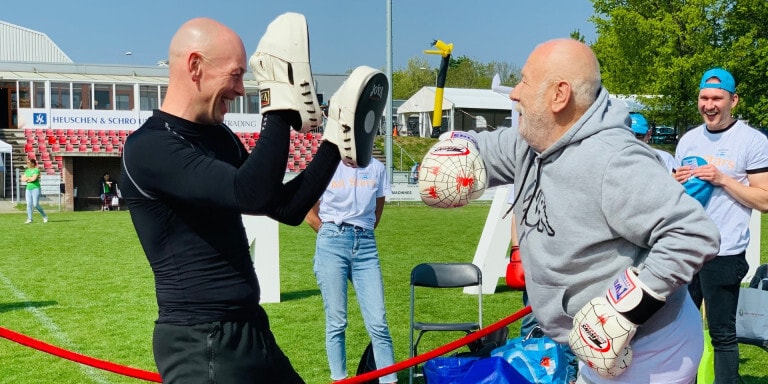Twee lachende mannen doen op een zonnig sportveld een speelse boksdemonstratie: de een draagt bokshandschoenen en de ander houdt stootkussens omhoog.
