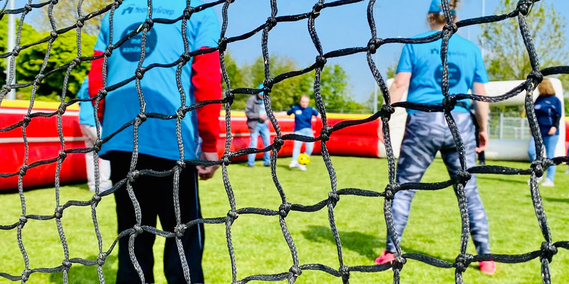In de voorgrond staat een doelnet en op de achtergrond spelen mensen voetbal op een grasveld met een opblaasbare boarding.
