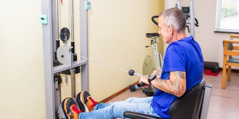 Een man in een blauw shirt zit op een fitnessapparaat en voert geconcentreerd een roeioefening uit in een trainingsruimte.