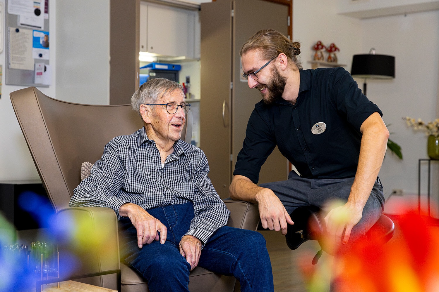 Een zorgmedewerker heeft een vrolijk en persoonlijk gesprek met een oudere man die comfortabel in een fauteuil zit in een huiselijke kamer.