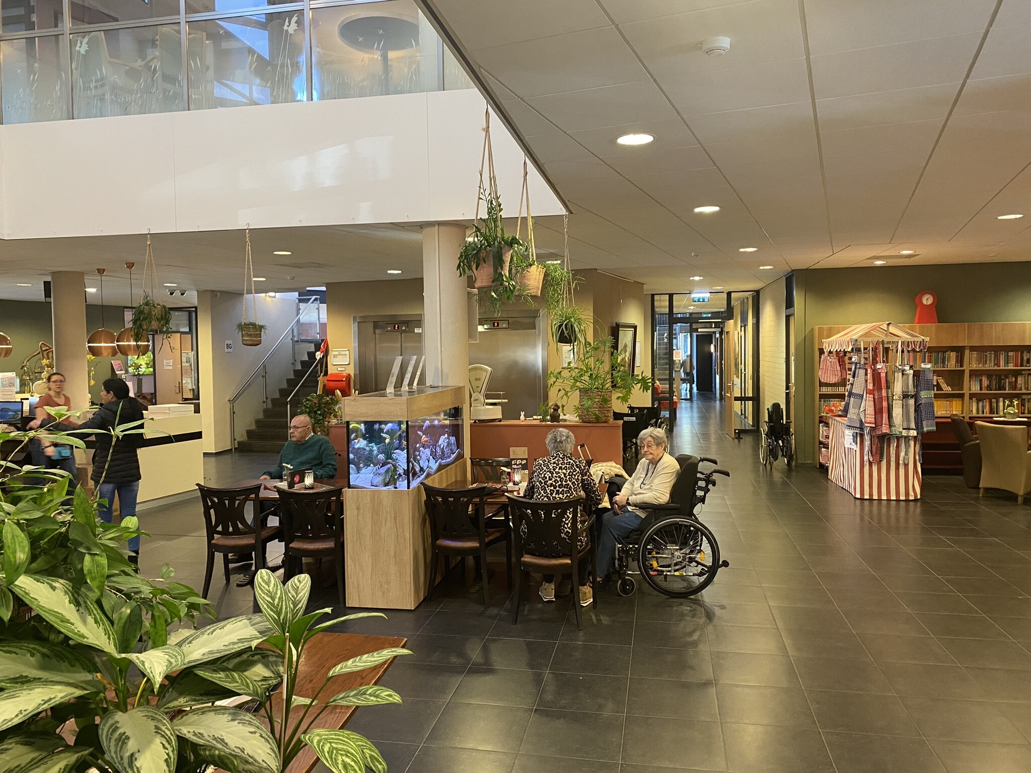 Een gezamelijke ruimte op de locatie Dokter Calshof. Aan een tafel zit een oude man, aan de andere tafel zitten twee oudere vrouwen waarvan een in een rolstoel.