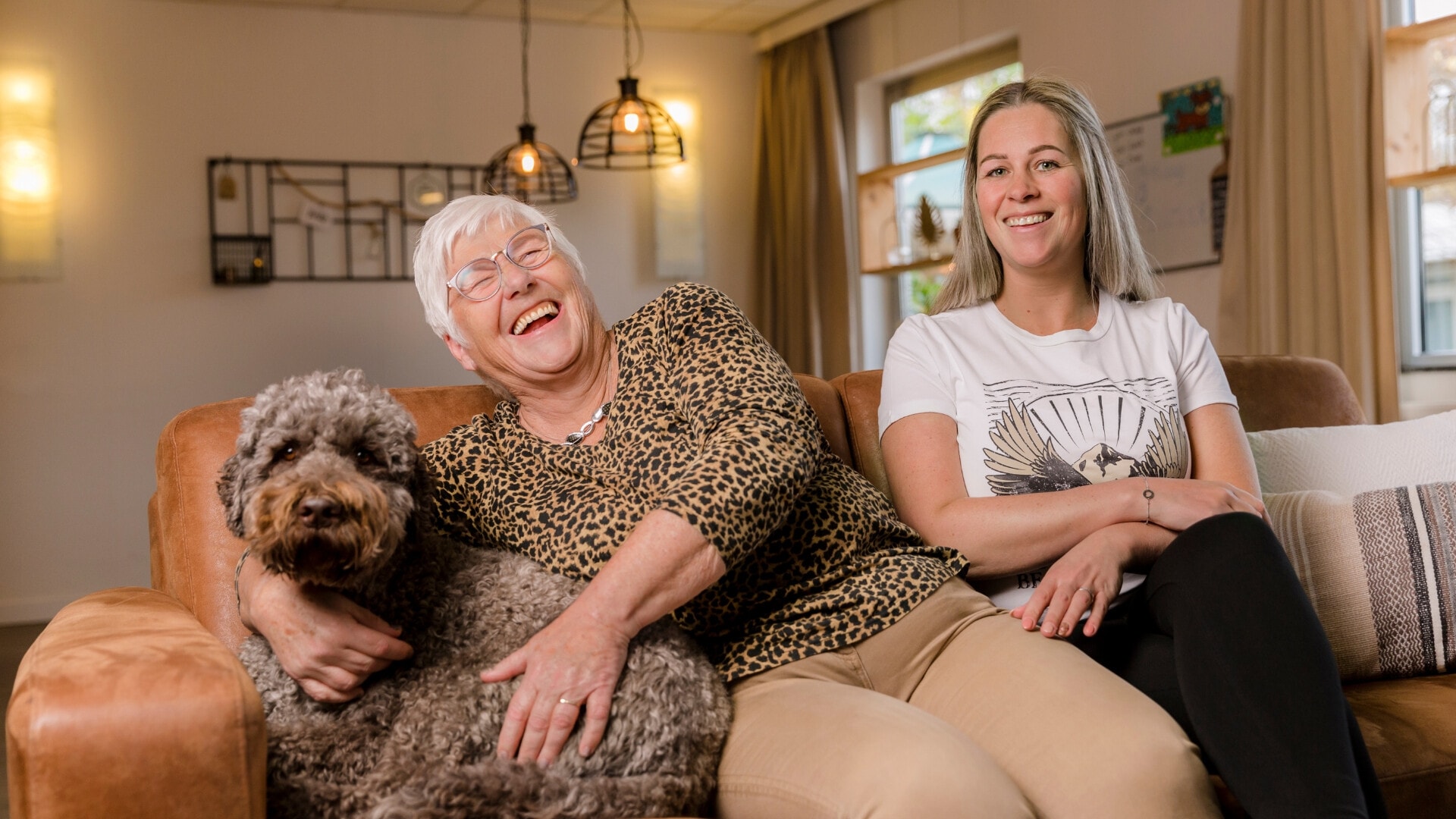 Lachende oude vrouw zit op de bank naast een jonge vrouw en een hond.