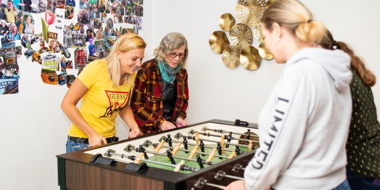 Vier vrouwen zijn aan het tafelvoetballen. Achter hen hangen veel foto's aan een witte muur.